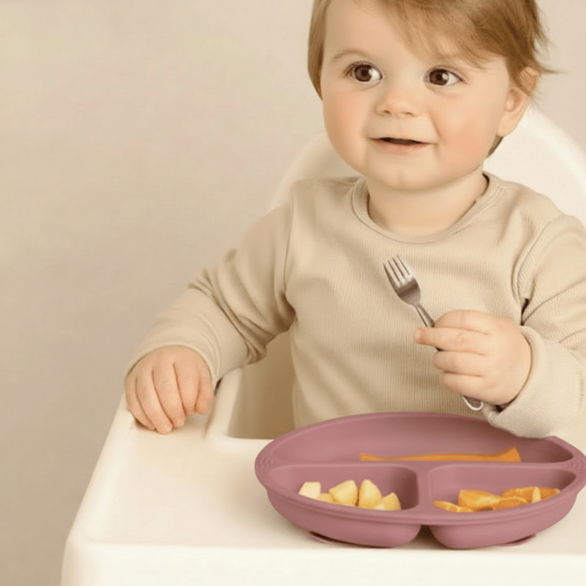 Un enfant prend ses aliments avec sa fourchette inox dans l'assiette bebe silicone  à 3 compartiments et ventouses coloris rose