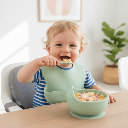 Un enfant installé sur un réhausseur de chaise mange des coquillettes au jambon dans son assiette bebe ventousée coloris vert avec sa cuillère assortie.