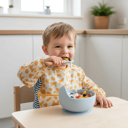 Un enfant portant un bavoir manches longues et assis à sa petite table mange un fromage blanc avec morceaux de fruits dans son assiette bebe ventousée coloris bleu et avec sa cuillère assortie.