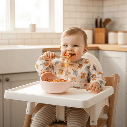 Une fillette portant un bavoir manches longues et installée dans sa chaise haute mange des spaghetti bolognaise dans son assiette bebe ventousée coloris rose avec sa cuillère assortie