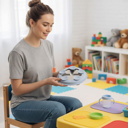 Une jeune maman assise sur une petite chaise d'enfant installe le couvert sur une petite table. Elle vérifie que le support bleu en silicone avec ses 5 ventouses soit bien ajusté à l'assiette bébé inox en la retournant.