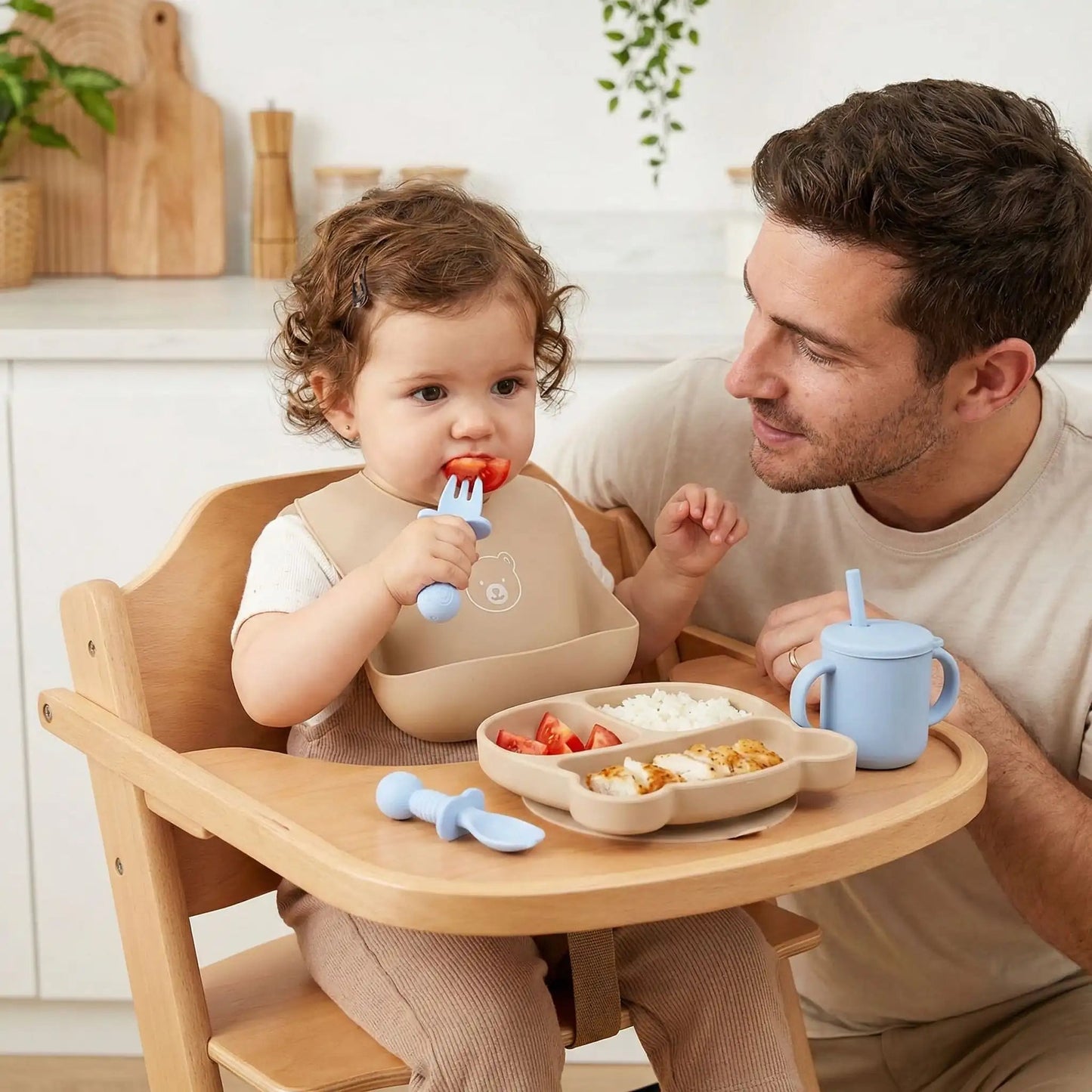 une petite fille mange un morceau de tomate avec son couvert bébé ergonomique bleu. Son papa est à ses côtés