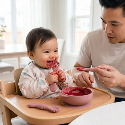 une fillette mange la puree de betterave contenue dans un bol ventousé à la tablette de chaise haute avec sa cuillere ergonomique bebe ajourée coloris rose. Elle est aidée par son père avec la cuillere ergonomique bebe pleine assortie.