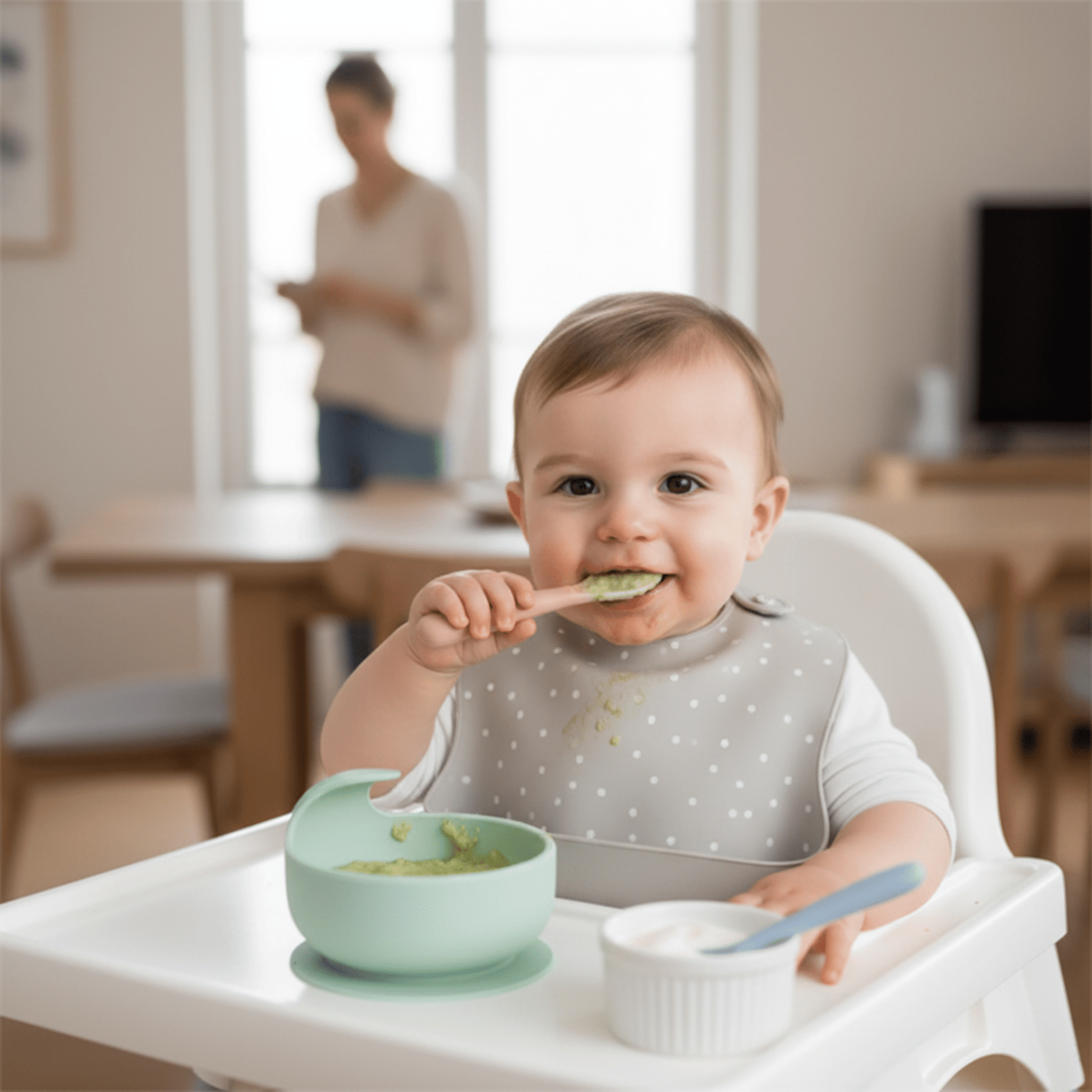 un enfant dans sa chaise haute utilise ses cuilleres bebe en silicone : coloris rose pour la purée de courgette et bleu pour le laitage, sous la surveilllance de sa maman