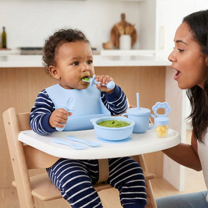 Un bébé de 8 mois amène à sa bouche la cuillère ergonomique silicone bleue contenant sa purée de légumes encouragé par sa mère qui ouvre également la bouche. Le bol de purée, la tasse à paille silicone bleue et la grignoteuse contenant de petits morceaux de fruits sont aussi sur la tablette de la chaise haute. Un repas équilibré en autonomie !