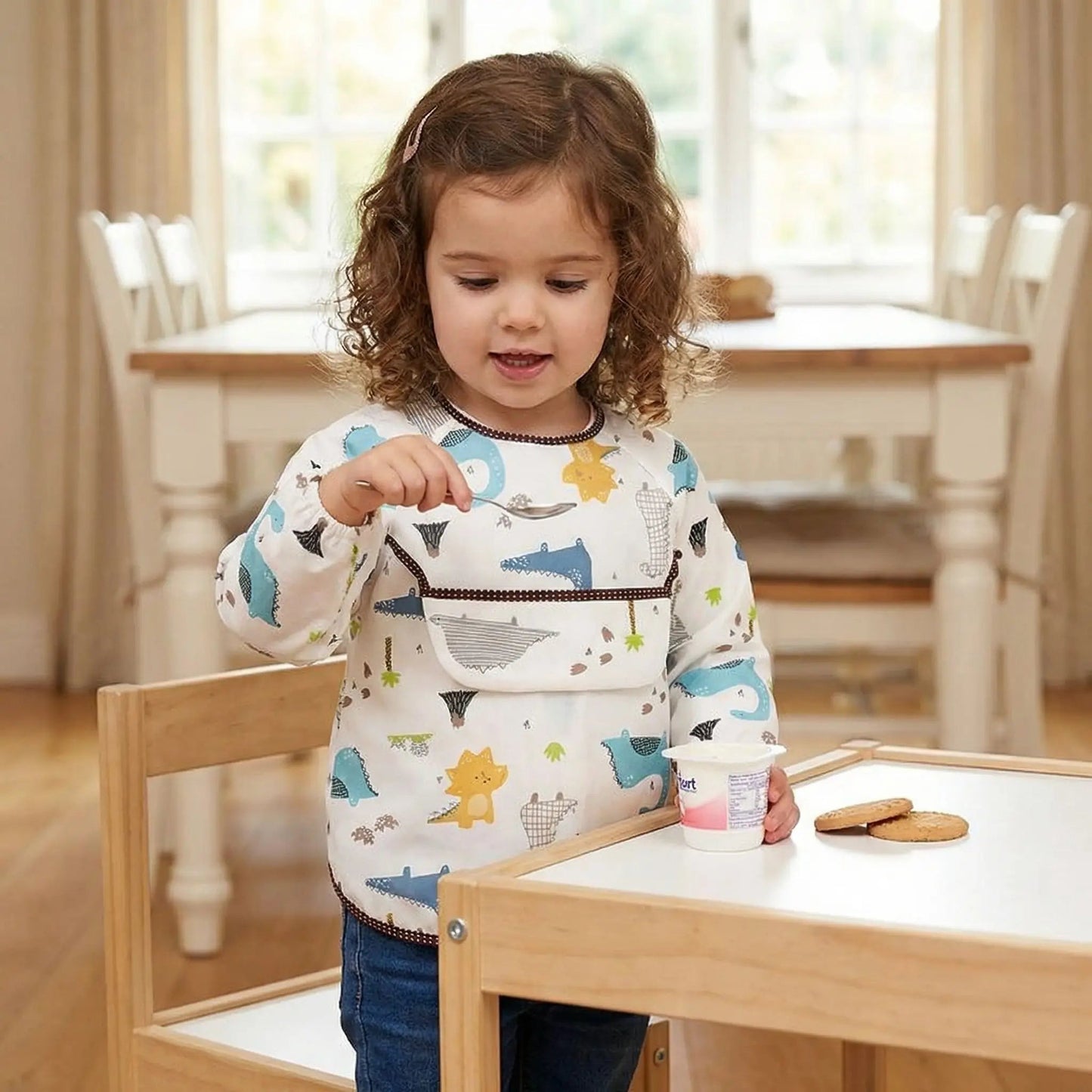 une petite fille se met debout pour manger son yaourt avec son bavoir manche longue imperméable modèle dinosaures