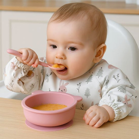 un petit enfant mange sa purée avec une cuillère dans son assiette pour bébé antidérapante ergonomique coloris rose