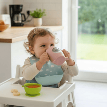fillette dans sa chaise haute devant une purée de carotte et des rondelles de banane boit dans sa tasse d'apprentissage bebe avec hublot rose clair