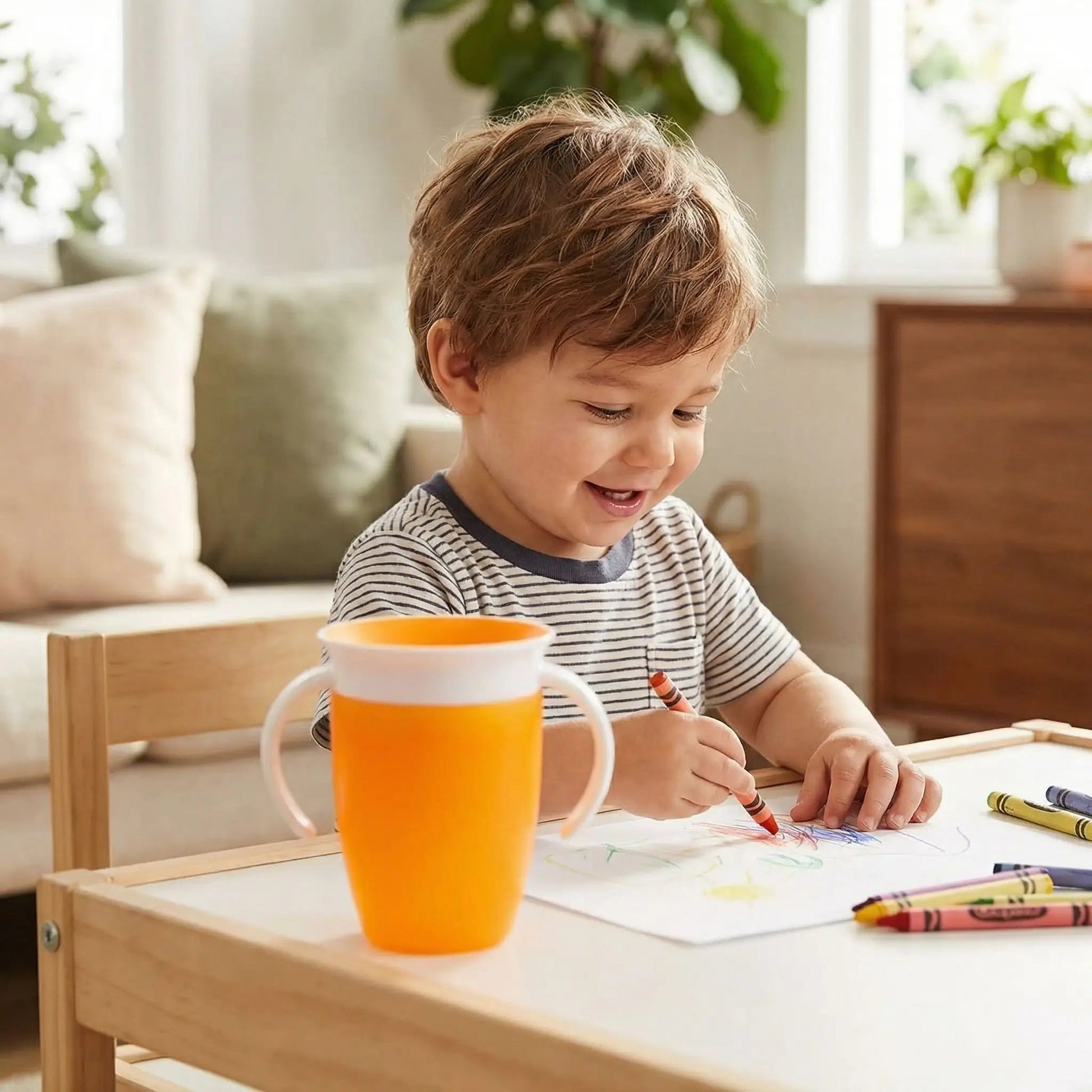 un enfant dessine à sa petite table sur laquelle est posée sa tasse d'apprentissage 360 coloris orange