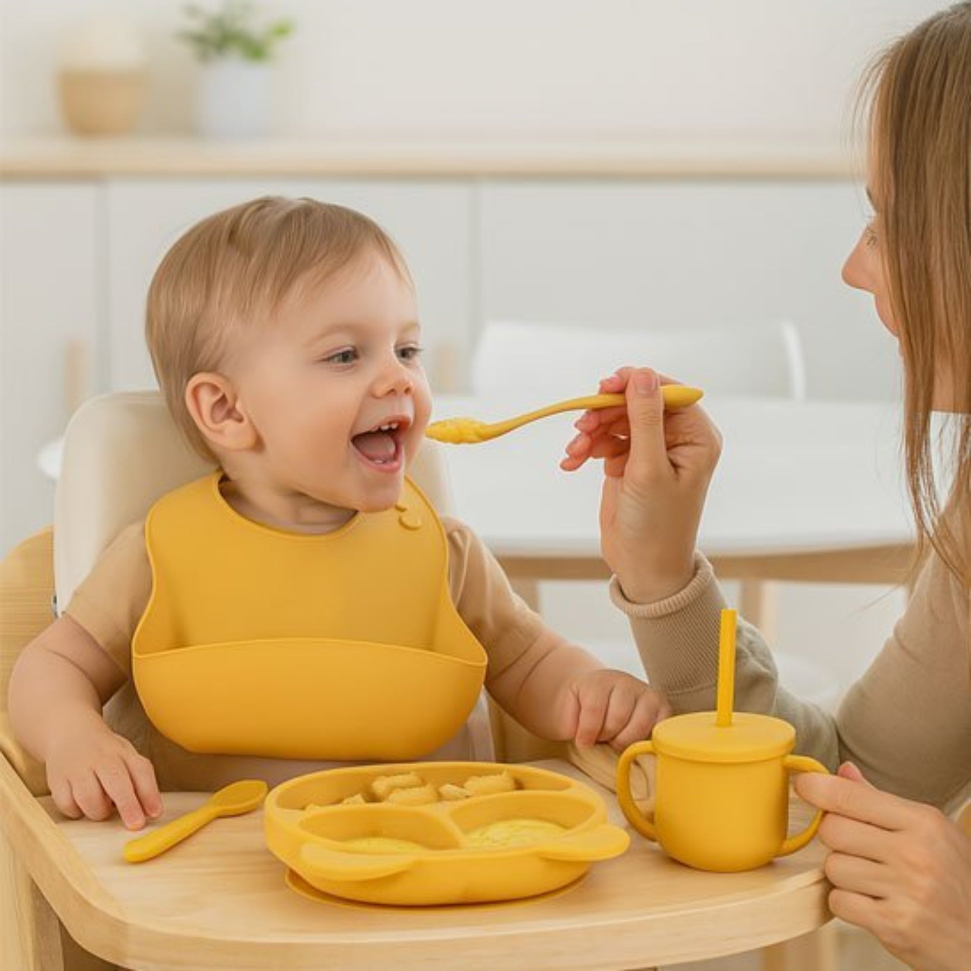 un petit garçon équipé du set vaisselle bébé silicone jaune ouvre grand sa bouche pour laisser sa mère lui donner la cuillère. Moment de partage.