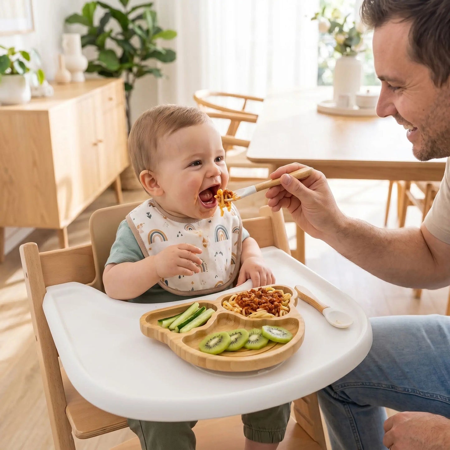 un papa nourrit son petit garçon avec la fourchette du set de vaisselle pour bebe coloris blanc. L'assiette compartimentée en bambou contient entrée, plat, dessert.